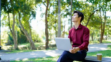 Parktaki bankta oturan bir adam yanında kahve fincanıyla dizüstü bilgisayar kullanarak güneşli bir günün tadını çıkarıyor..
