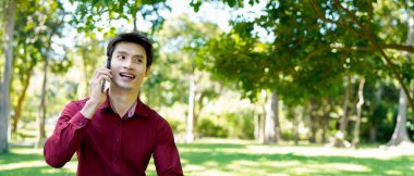 Parkta yürürken akıllı telefon kullanan mutlu Asyalı bir adam güneşli bir günde, yemyeşil ve modern mimariyle çevrili güzel bir sohbetin tadını çıkarıyor..