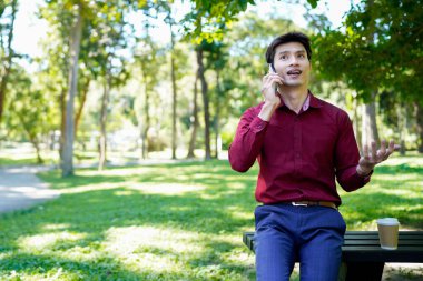 Parkta akıllı telefon kullanan mutlu Asyalı bir adam, güneşli bir günde keyifli bir sohbetin tadını çıkarıyor, yemyeşil ve modern mimariyle çevrili...