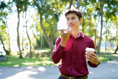 Paket kahveli genç Asyalı adam şehir parkında akıllı telefondan iletişim kuruyor..