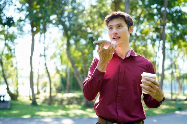 Paket kahveli genç Asyalı adam şehir parkında akıllı telefondan iletişim kuruyor..