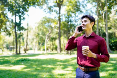 Paket kahveli genç Asyalı adam şehir parkında akıllı telefondan iletişim kuruyor..