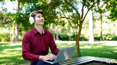 Asian man smiling, enjoying remote work outdoors, typing on a laptop and listening to music through headphones in a park setting.