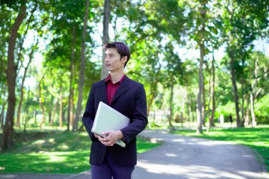 Siyah takım elbiseli profesyonel Asyalı işadamı parkta dizüstü bilgisayar taşıyor..