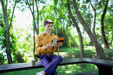 Genç adam parktaki bankta eğleniyor kitap okuyor ve güneşli bir günde kahve içiyor..