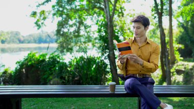 Genç adam parktaki bankta eğleniyor kitap okuyor ve güneşli bir günde kahve içiyor..