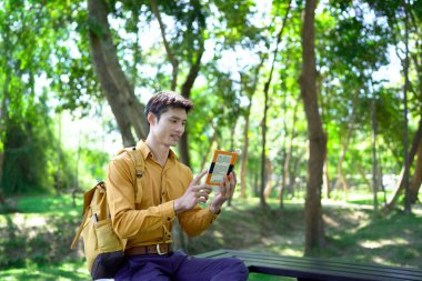 Genç adam parktaki bankta eğleniyor kitap okuyor ve güneşli bir günde kahve içiyor..