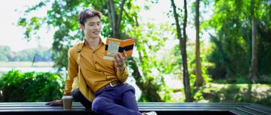 Genç adam parktaki bankta eğleniyor kitap okuyor ve güneşli bir günde kahve içiyor..