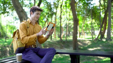 Genç adam parktaki bankta eğleniyor kitap okuyor ve güneşli bir günde kahve içiyor..