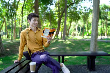 Genç adam parktaki bankta eğleniyor kitap okuyor ve güneşli bir günde kahve içiyor..