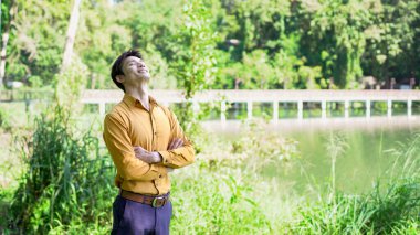Genç Asyalı bir adam parkta gözleri kapalı dinleniyor. Huzurlu bir adam doğanın tadını çıkarıyor ve dikkat çekmek ve stres atmak için temiz hava soluyor...