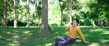 Parkta akıllı telefon kullanan mutlu Asyalı bir adam, güneşli bir günde keyifli bir sohbetin tadını çıkarıyor, yemyeşil ve modern mimariyle çevrili...