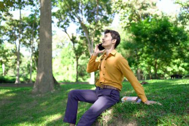 Parkta akıllı telefon kullanan mutlu Asyalı bir adam, güneşli bir günde keyifli bir sohbetin tadını çıkarıyor, yemyeşil ve modern mimariyle çevrili...