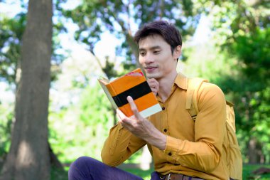 Erkek, güneşli bir günde, sakin bir parkta, büyük bir ağacın altında kitap okumaktan hoşlanır..