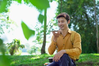 Parkta akıllı telefon kullanan mutlu Asyalı bir adam, güneşli bir günde keyifli bir sohbetin tadını çıkarıyor, yemyeşil ve modern mimariyle çevrili...