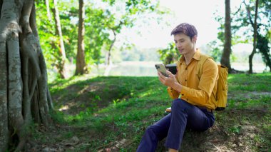 Paket kahveli genç Asyalı adam şehir parkında akıllı telefondan iletişim kuruyor..