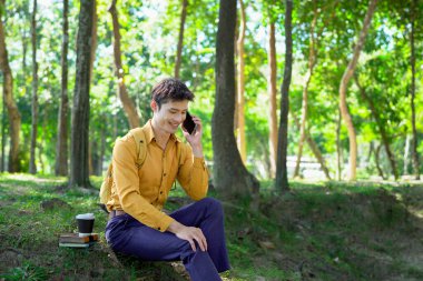 Parkta akıllı telefon kullanan mutlu Asyalı bir adam, güneşli bir günde keyifli bir sohbetin tadını çıkarıyor, yemyeşil ve modern mimariyle çevrili...