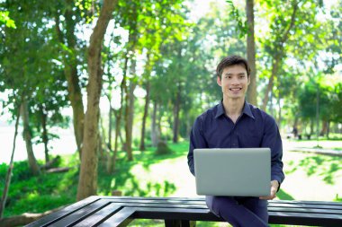 Genç adam güneşli bir park ortamında dizüstü bilgisayarda çalışırken aynı zamanda canlı ve profesyonel bir yaşam tarzının tadını çıkarıyor..