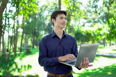 Genç adam güneşli bir park ortamında dizüstü bilgisayarda çalışırken aynı zamanda canlı ve profesyonel bir yaşam tarzının tadını çıkarıyor..