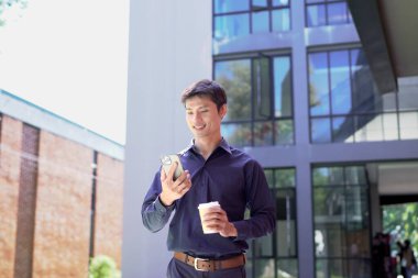 Gülümseyen işadamı akıllı telefon kullanıyor ve ofis binasının dışında kahve tutuyor..