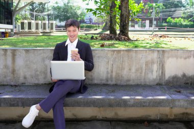 Parkta dizüstü bilgisayarda çalışan, parktaki bankta kahve içen genç bir adam..