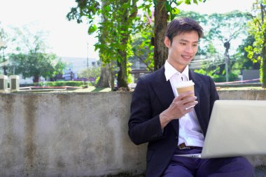 Parkta dizüstü bilgisayarda çalışan, parktaki bankta kahve içen genç bir adam..