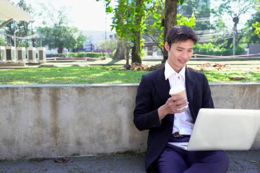 Parkta dizüstü bilgisayarda çalışan, parktaki bankta kahve içen genç bir adam..