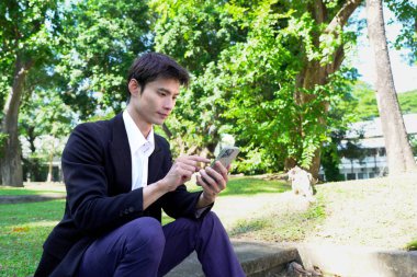 Genç Asyalı işadamı parkta akıllı telefon kullanıyor..