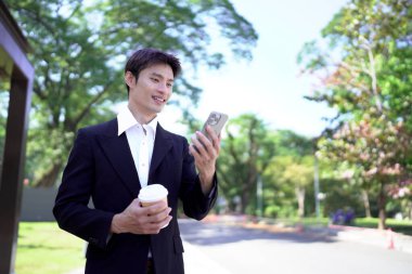 Siyah takım elbiseli genç Asyalı işadamı elinde hazır kahveyle şehir parkında akıllı telefon kullanıyor..