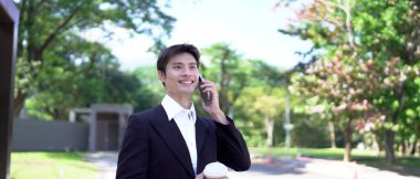 Siyah takım elbiseli genç Asyalı işadamı şehir parkında akıllı telefondan iletişim kuruyor...