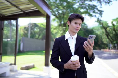 Siyah takım elbiseli genç Asyalı işadamı elinde hazır kahveyle şehir parkında akıllı telefon kullanıyor..