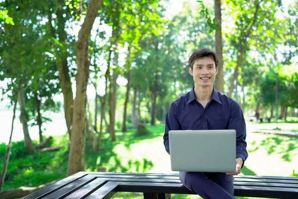 Genç adam güneşli bir park ortamında dizüstü bilgisayarda çalışırken aynı zamanda canlı ve profesyonel bir yaşam tarzının tadını çıkarıyor..