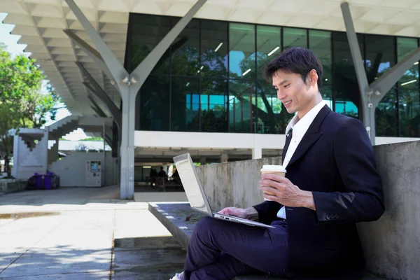 Kentsel bankta oturan iş adamı, dizüstü bilgisayarla çalışıyor ve cam mimarinin önünde kahve fincanı tutuyor..