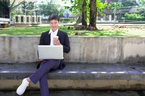 Parkta dizüstü bilgisayarda çalışan, parktaki bankta kahve içen genç bir adam..