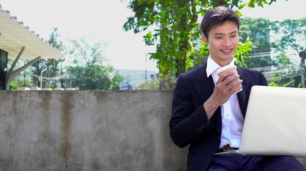 Parkta dizüstü bilgisayarda çalışan, parktaki bankta kahve içen genç bir adam..