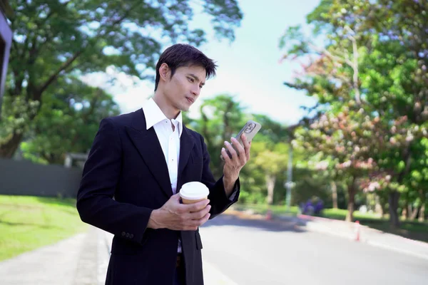 Siyah takım elbiseli genç Asyalı işadamı elinde hazır kahveyle şehir parkında akıllı telefon kullanıyor..