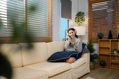 Huzurlu genç Asyalı kadın kablosuz kulaklıkla müzik dinlerken oturma odasındaki kanepede rahatça oturuyor, akıllı telefon tutuyor ve rahat ev ortamında dinleniyor..