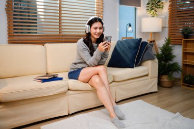 Huzurlu genç Asyalı kadın kablosuz kulaklıkla müzik dinlerken oturma odasındaki kanepede rahatça oturuyor, akıllı telefon tutuyor ve rahat ev ortamında dinleniyor..