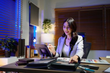 İş yerinde İç Tasarımcı: Asyalı Yetişkin Kadın Gece Masasında Mimari Planlarla Ev Yenileme Projesi için malzeme örnekleri ve renk örnekleri seçiyor.