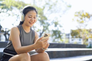 Young female athlete rests between workouts, sitting relaxed on the steps and communicating or making selfie using smartphone. Attractive fitness girl in gray sportswear relaxes after jogging..