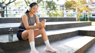 Genç bayan sporcu idmanlar arasında dinleniyor, basamaklarda rahatça oturuyor ve akıllı telefon kullanarak iletişim kuruyor ya da selfie çekiyor. Gri spor kıyafetli çekici fitness kızı koşudan sonra rahatlıyor...