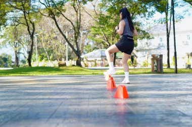 Bir kadın turuncu koni ile parkta egzersiz yapıyor. Külahlar sıraya dizilmiş ve kadın onların arasından koşuyor..