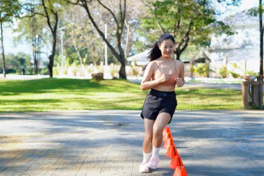 Bir kadın turuncu koni ile parkta egzersiz yapıyor. Külahlar sıraya dizilmiş ve kadın onların arasından koşuyor..