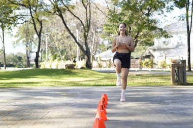 Bir kadın turuncu koni ile parkta egzersiz yapıyor. Külahlar sıraya dizilmiş ve kadın onların arasından koşuyor..