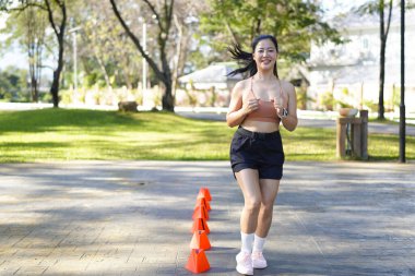 Bir kadın turuncu koni ile parkta egzersiz yapıyor. Külahlar sıraya dizilmiş ve kadın onların arasından koşuyor..