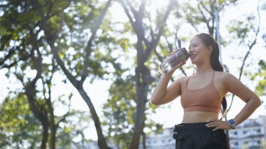 Genç Asyalı kadın parkta egzersiz yaparken şişeden su içiyor..