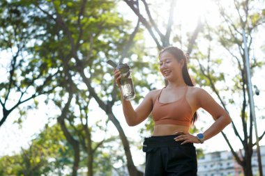 Genç Asyalı kadın parkta egzersiz yaparken şişeden su içiyor..