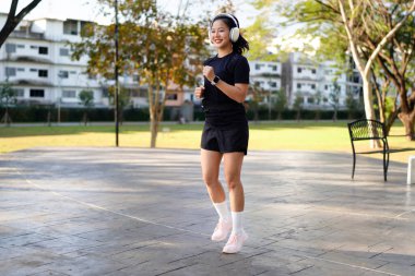 Kulaklık takan genç Asyalı kadın vücut boyu dışarıda antrenman yapıyor, ısınıyor ve yükseğe zıplıyor, spor kıyafetli kadın dışarıda antrenman yapmaktan hoşlanıyor..