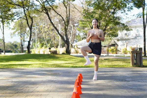 Bir kadın turuncu koni ile parkta egzersiz yapıyor. Külahlar sıraya dizilmiş ve kadın onların arasından koşuyor..