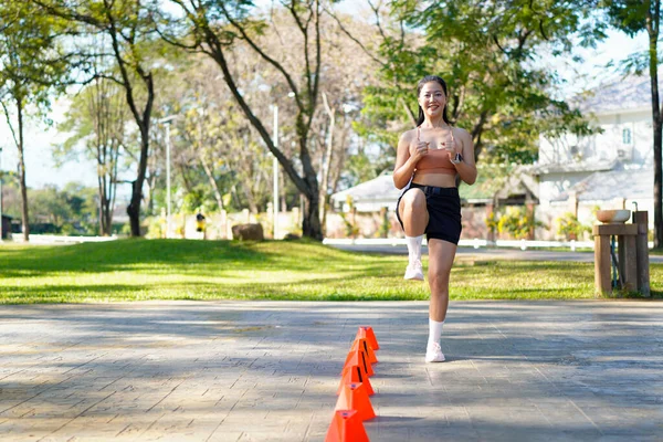 Bir kadın turuncu koni ile parkta egzersiz yapıyor. Külahlar sıraya dizilmiş ve kadın onların arasından koşuyor..